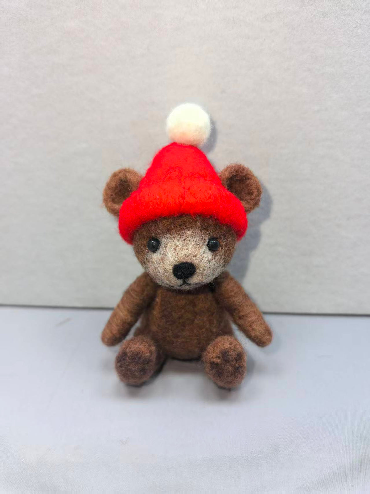 Needle felted brown teddy bear wearing a red beanie with a white pom-pom