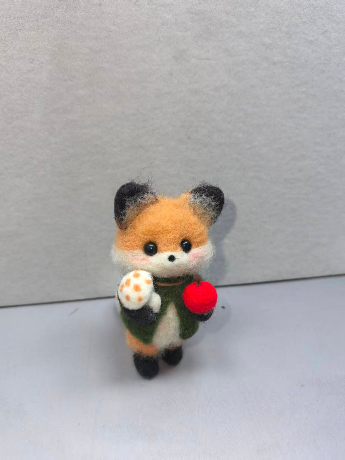 Needle felted fox in a green vest holding a spotted mushroom and a red apple