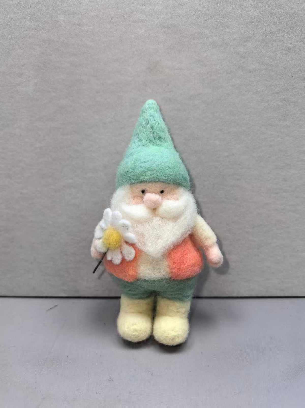 Needle felted gnome in a mint hat and coral vest holding a daisy