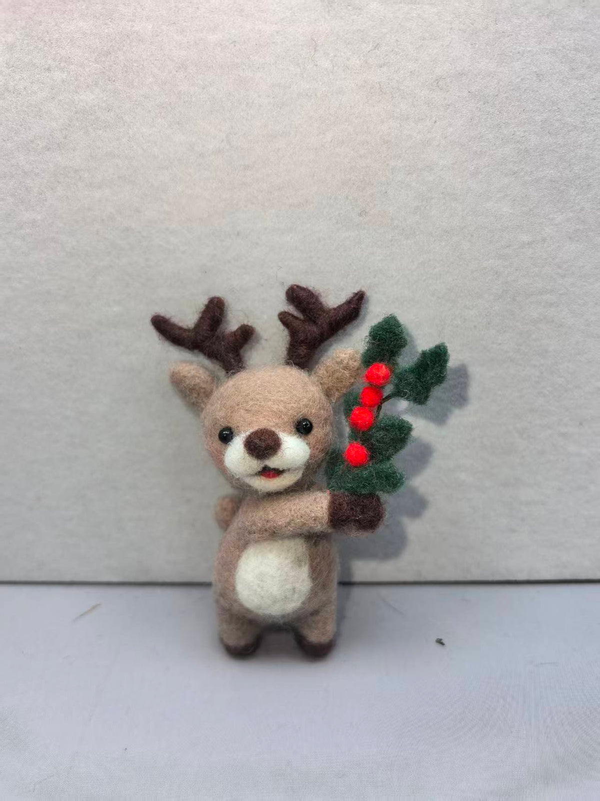 Needle felted reindeer with brown antlers holding a holly sprig with red berries